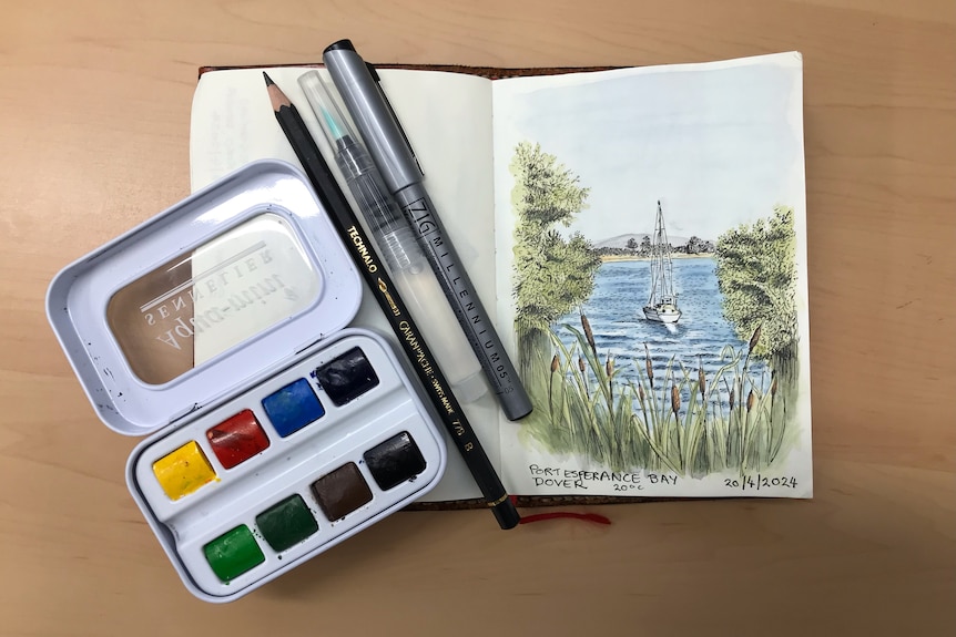 A small book open to a picture of a boat. Paints and pencils on the left hand side.