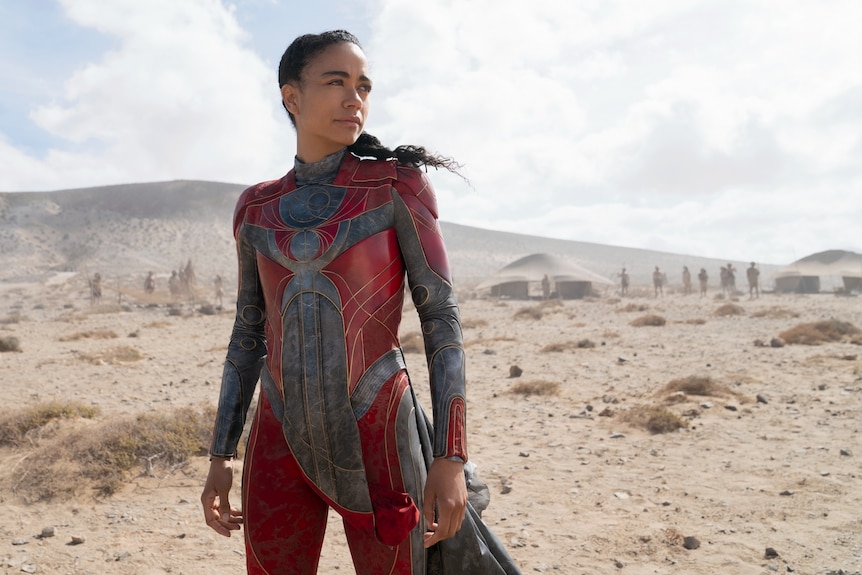 Lauren Ridloff, in a red superhero costume, strikes a resolute pose, staring off into the distance in the desert