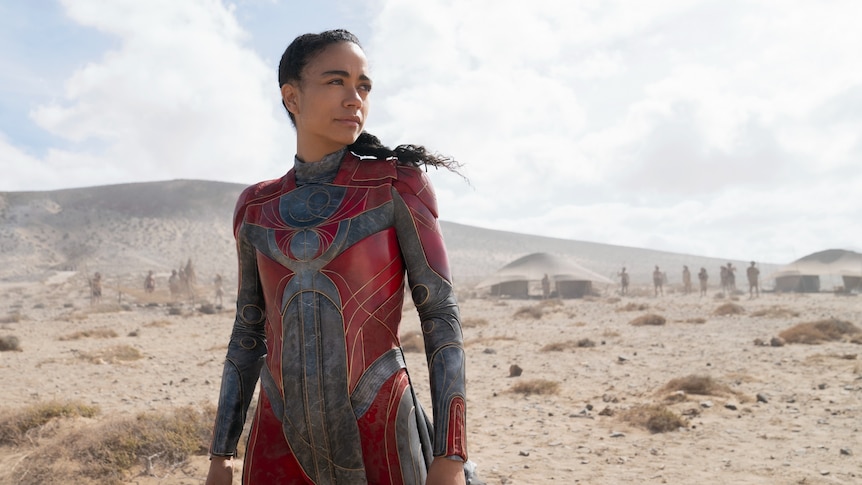 Lauren Ridloff, in a red superhero costume, strikes a resolute pose, staring off into the distance in the desert