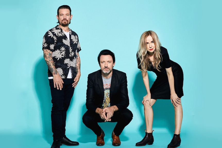 Three members of a band crouch and stand in front of a light blue backdrop