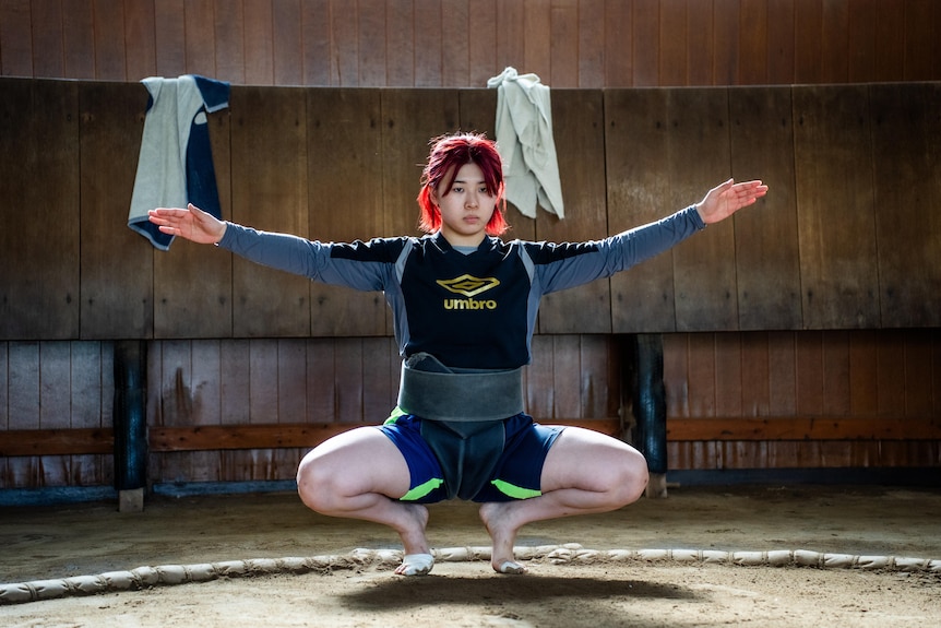 A woman with red hair squats with her arms oustretched 
