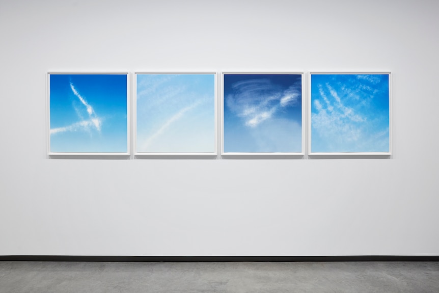 Four artworks hanging on a white gallery wall showing white clouds against blue sky.
