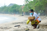 An older Balinese women crouches on a beach holding a woven basket