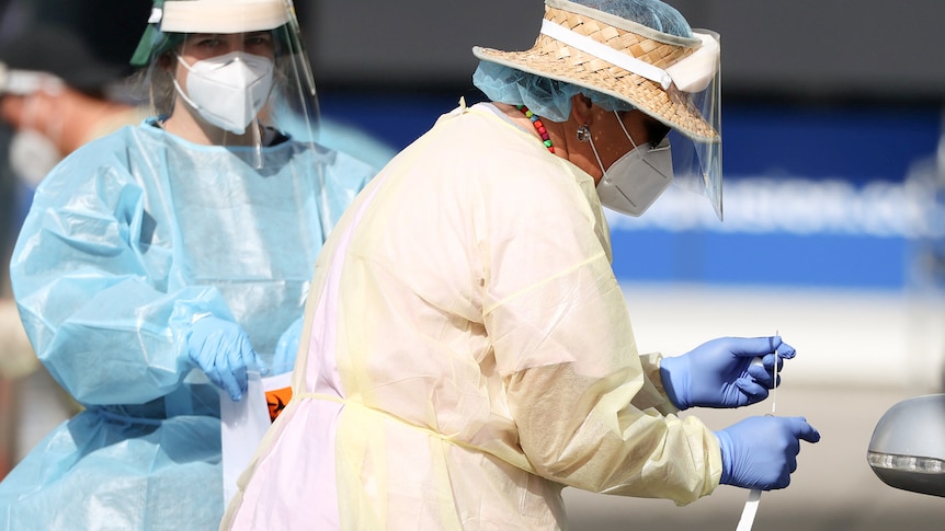 A woman wearing a face shield and mask and gloves looks down at a COVID swab test in her hand.