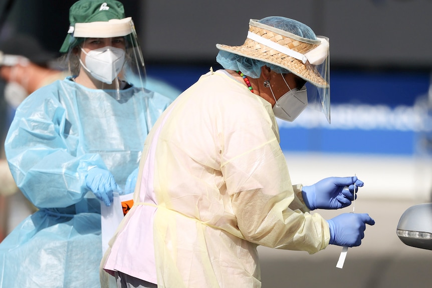 A woman wearing a face shield and mask and gloves looks down at a COVID swab test in her hand.