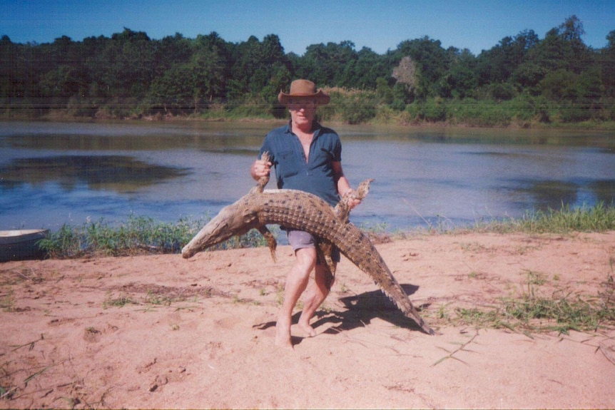 An older Bill Dean holds a crocodile by a river bank