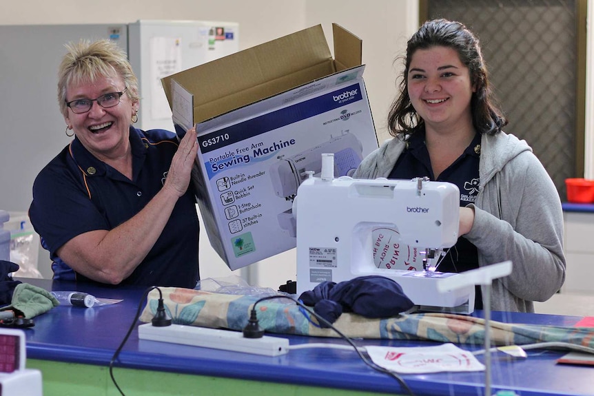 A Girl Guide leader holds an empty sewing machine box aloft while a Girl Guide sews beside her.