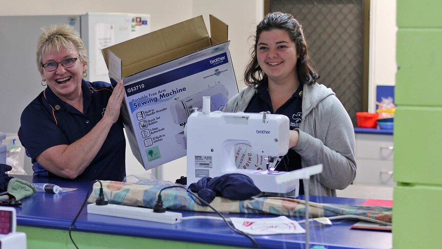 A Girl Guide leader holds an empty sewing machine box aloft while a Girl Guide sews beside her.