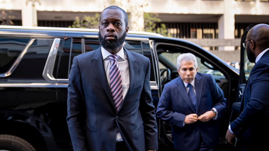 Black man with very short hair and beard, unsmiling, after just stepping out of black car. He is wearing a dark suit and tie