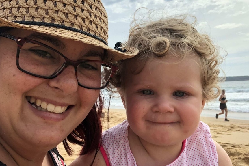 A bespectacled woman in a hat on the beach with a small, fair-haired child, smiling.