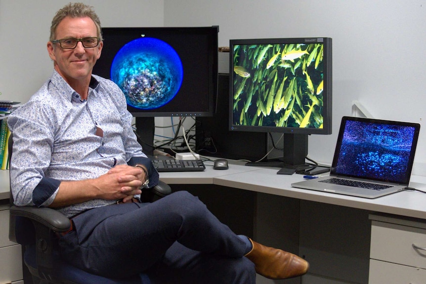 Man sitting at a desk with three computer monitors with photos of the reef on them.