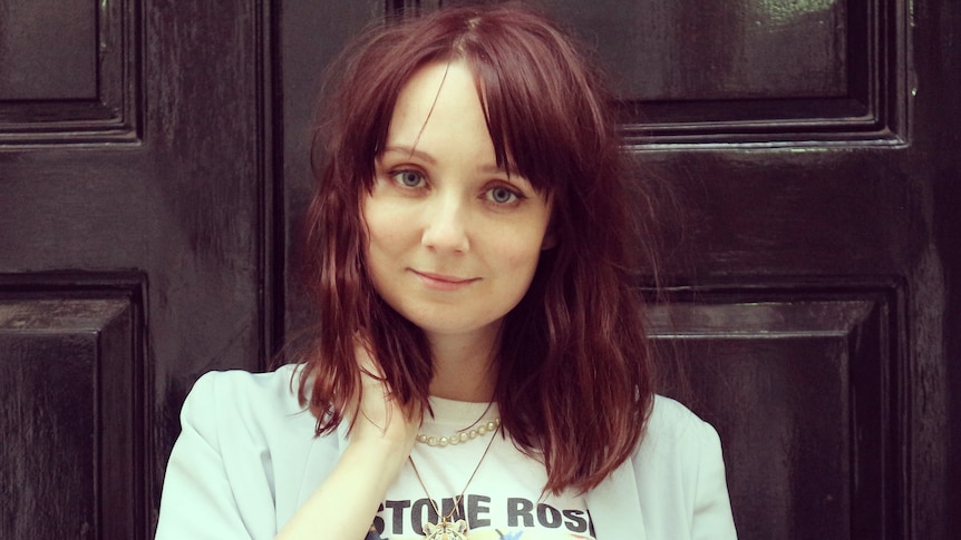 Claire Eastham, with shoulder-length brown hair, smiles standing in front of a large brown door.