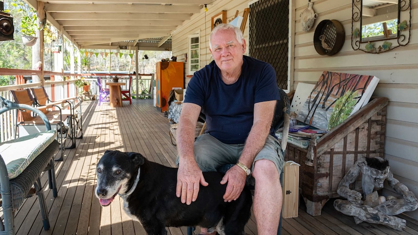 Wide shot of man seated in the foreground patting a medium sized dog, on a Queensland style verandah
