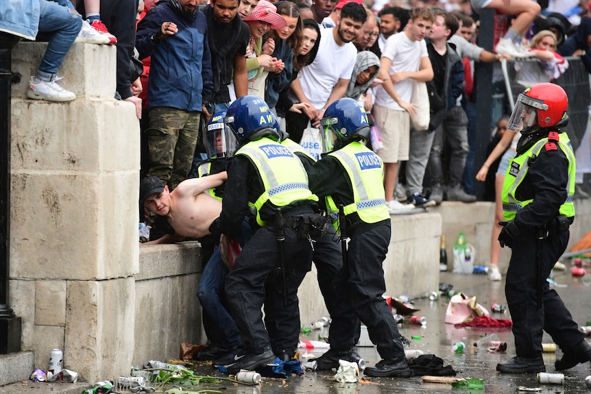 A shirtless man is detained by two police officers as a crowd of people watch on