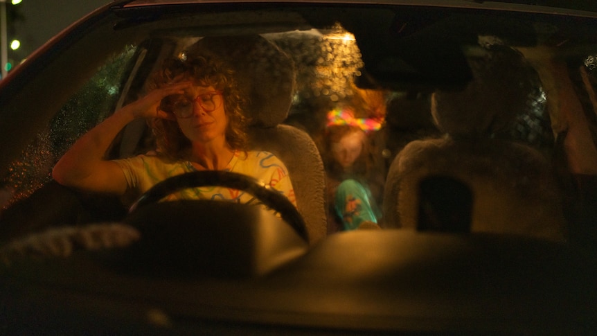 A woman is seen through the windshield of a car at night. She has her hands on the wheel. A child is in the back seat.