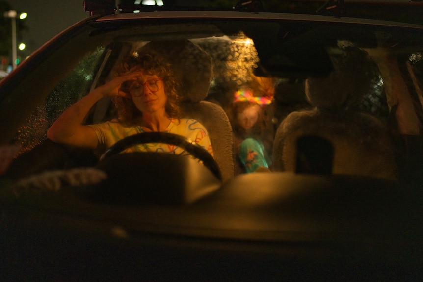 A woman is seen through the windshield of a car at night. She has her hands on the wheel. A child is in the back seat.