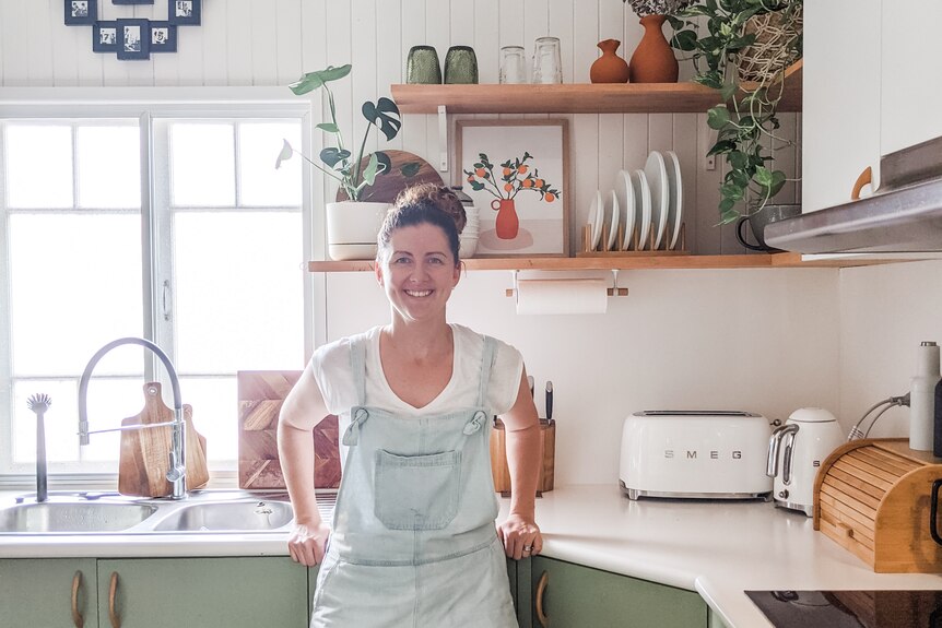 A woman in overalls leans against a green kitchen bench smiling.