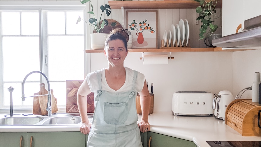 A woman in overalls leans against a green kitchen bench smiling.