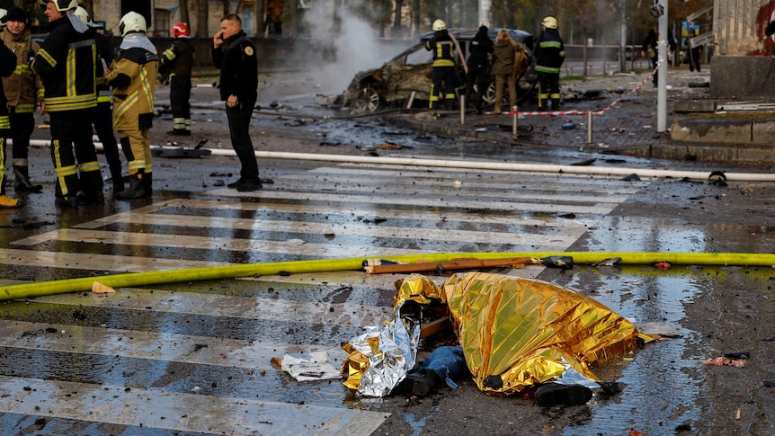 The legs of a dead body are visible underneath a yellow reflective blanket lying on a destroyed city street.