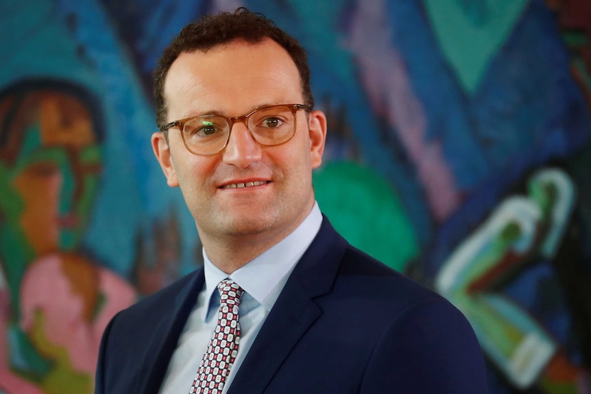 A mid-shot of Jens Spahn shows the minister in a blue suit, standing in front of a multi-coloured artwork.
