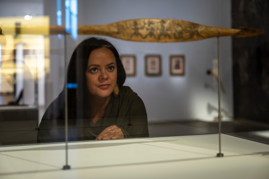 Woman with long dark hair and brown eyes wears an olive green dress and peers curiously into a cabinet featuring Indigenous art.