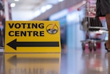 A yellow sign featuring the words 'voting centre' in black, with the feet of a man pushing a trolley in the background.