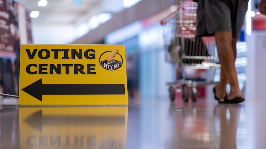 A yellow sign featuring the words 'voting centre' in black, with the feet of a man pushing a trolley in the background.