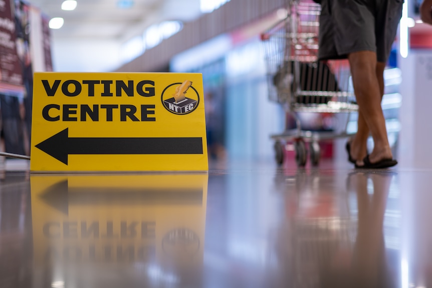 A yellow sign featuring the words 'voting centre' in black, with the feet of a man pushing a trolley in the background.