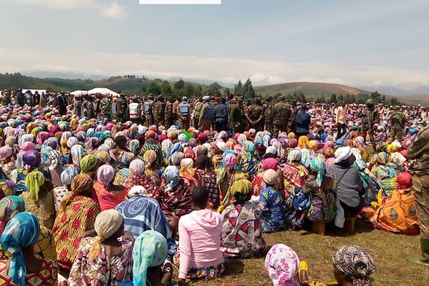 Hundreds of women sit on the ground outside wearing colourful clothes while soldiers look on