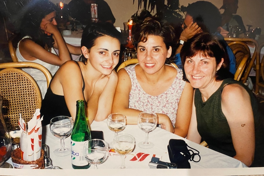 Three women, one in her 20s, one in her 30s, and one middle-aged, sit close together at a dining table, smiling widely