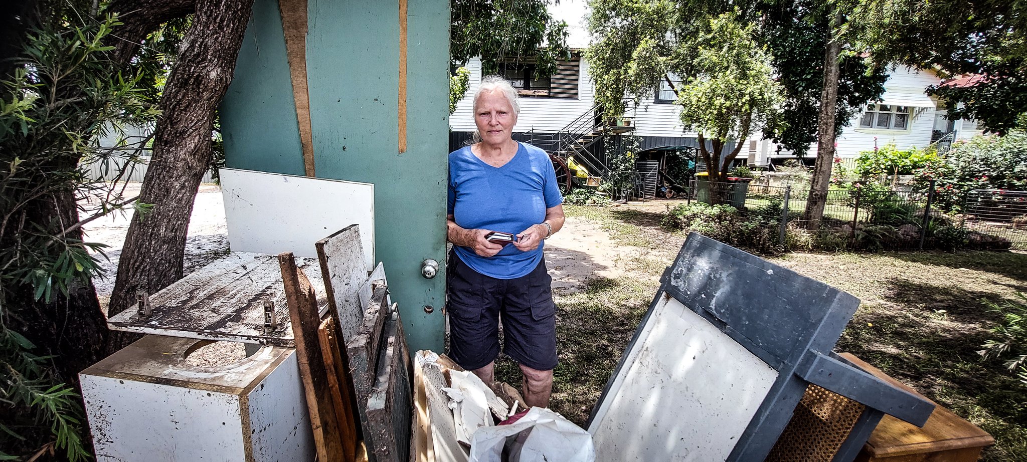 A 75-year-old woman standing in the front yard of a home behind a pile of debris from the flood.
