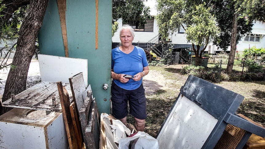 A 75-year-old woman standing in the front yard of a home behind a pile of debris from the flood.