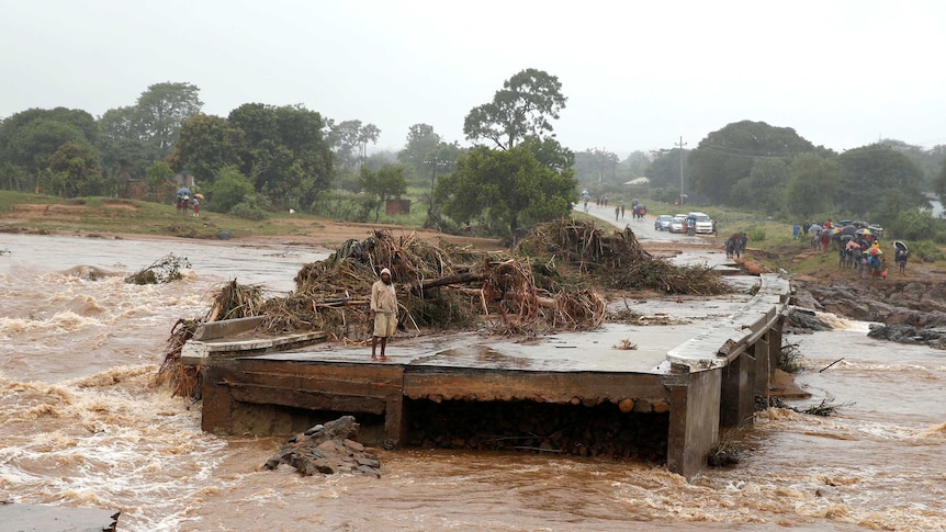 From a riverbank, a brown torrent races from left to right, with a man stands on a bridge that has been partly washed away.