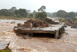 From a riverbank, a brown torrent races from left to right, with a man stands on a bridge that has been partly washed away.