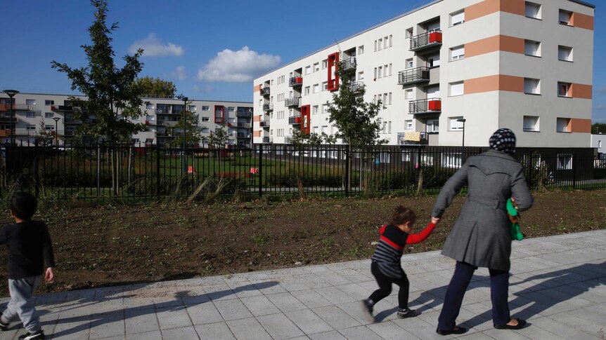 A woman holds her child's hand as they walk through a renovated area of Denain. a white unit complex is in the background.