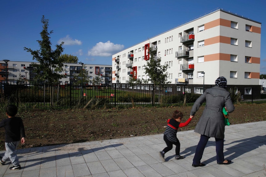 A woman holds her child's hand as they walk through a renovated area of Denain. a white unit complex is in the background.