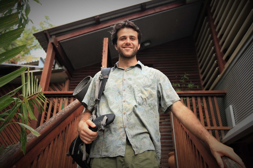 Sayle Johnston is wearing a backpack and walking down the steps of a wooden verandah. He is smiling.