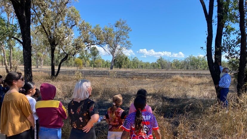 A group of students and teachers learn from an Indigenous man pointing at a burnt tree