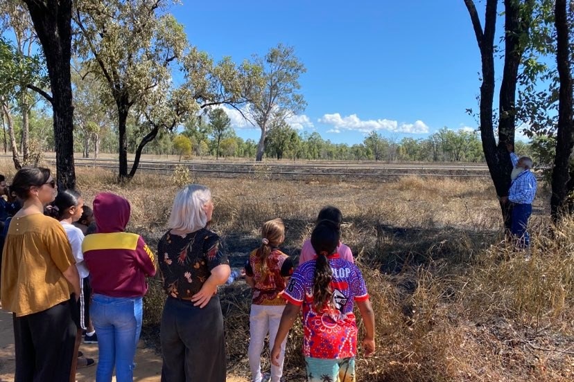 A group of students and teachers learn from an Indigenous man pointing at a burnt tree