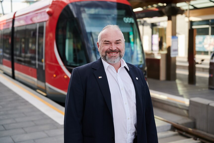A man with grey hair wearing a suit smiles in front of a red tram