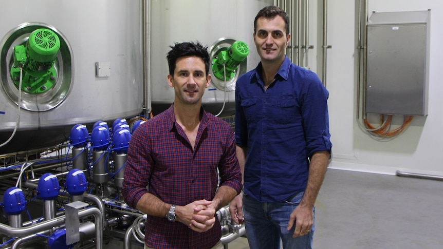 Luke and Matt stand before a series of large stainless steel tanks in an otherwise sterile room