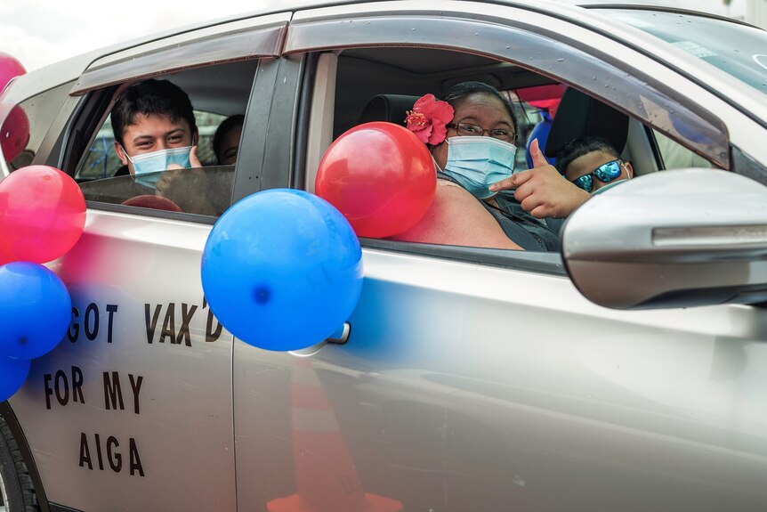 A woman and man smile out a car window. The car has balloons and the words 'I got vax'd for my aiga' on the side. 