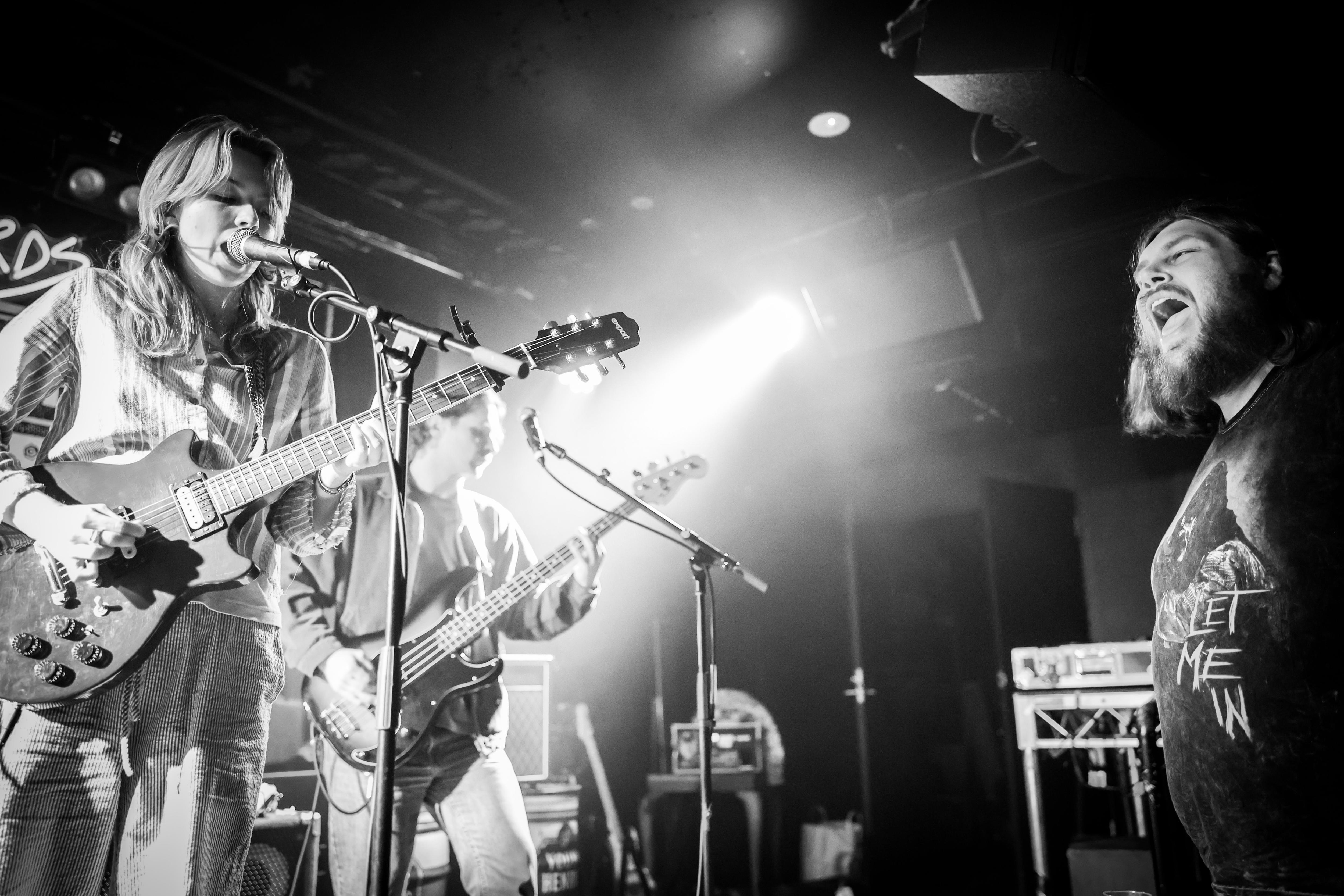 A man in his thirties with long hair and a beard watches two women onstage playing bass and guitar