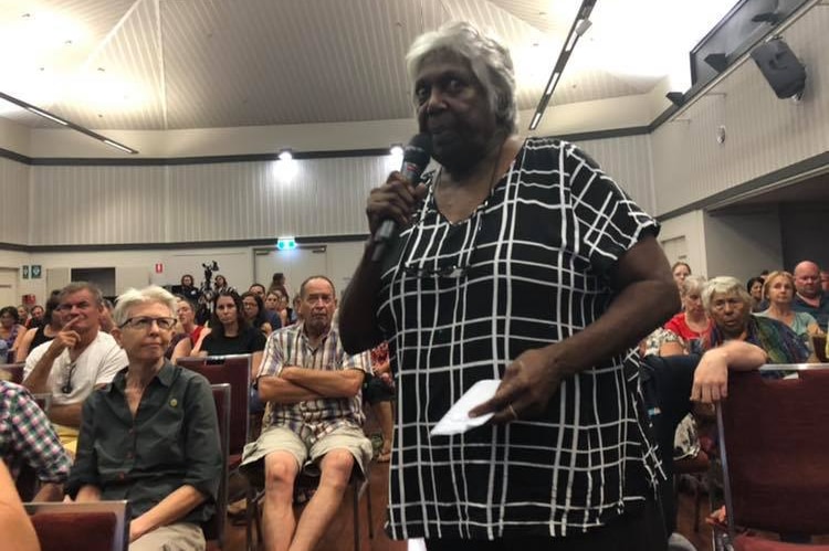 An Indigenous woman standing and speaking into a microphone, with the audience behind her.