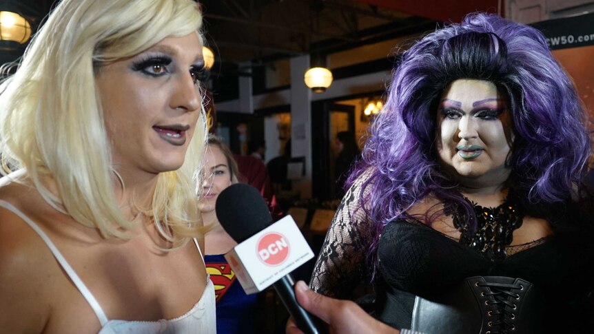 Blonde drag queen talking into a microphone (L) standing next to a drag queen with a purple wig