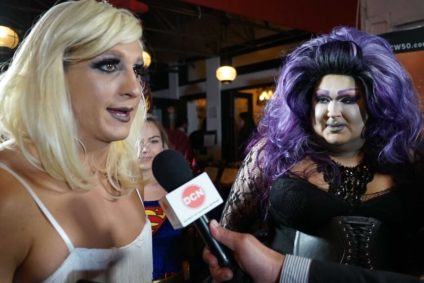 Blonde drag queen talking into a microphone (L) standing next to a drag queen with a purple wig
