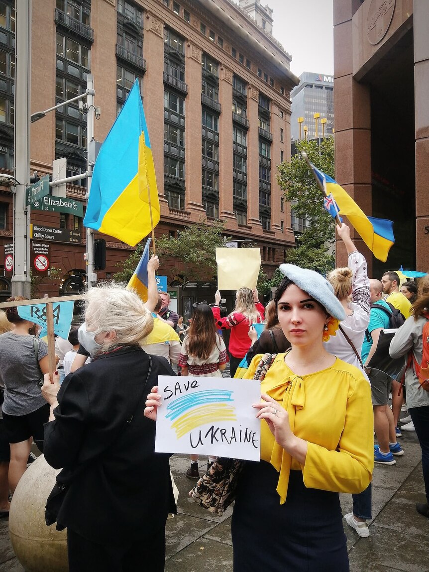 A young woman in a beret-style hat and a bright yellow blouse holds a sign saying "Save Ukraine" at a street rally.