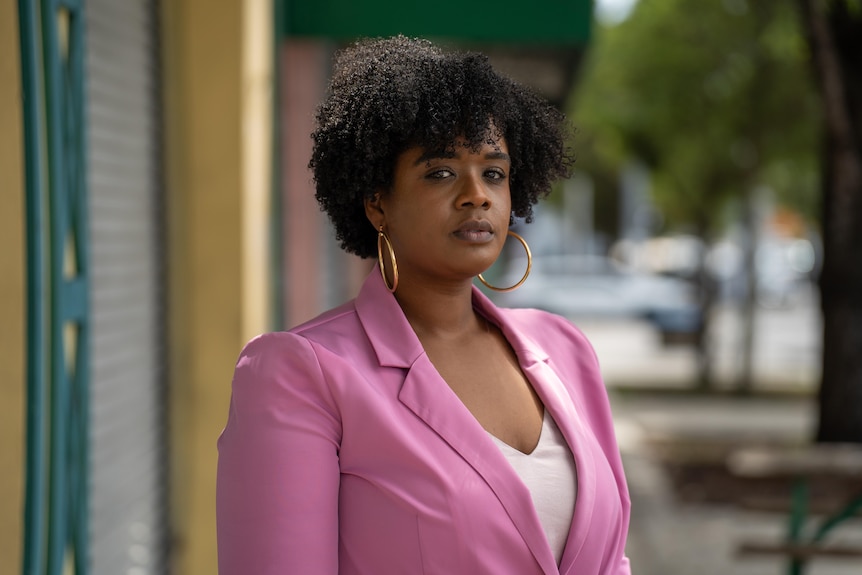 A Black woman wearing hoop earrings and a pink blazer stands outside.
