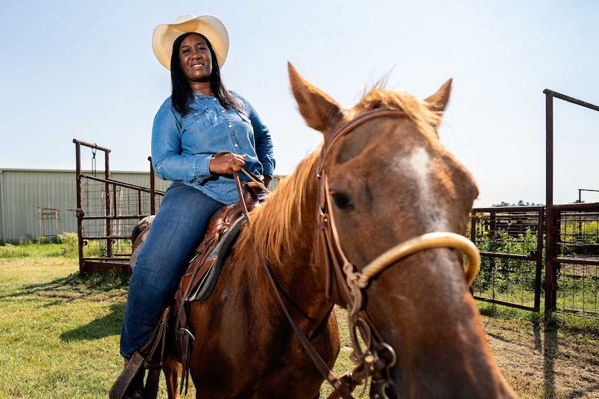 A woman in jeans, a denim shirt and cowboy hat sitting on a horse
