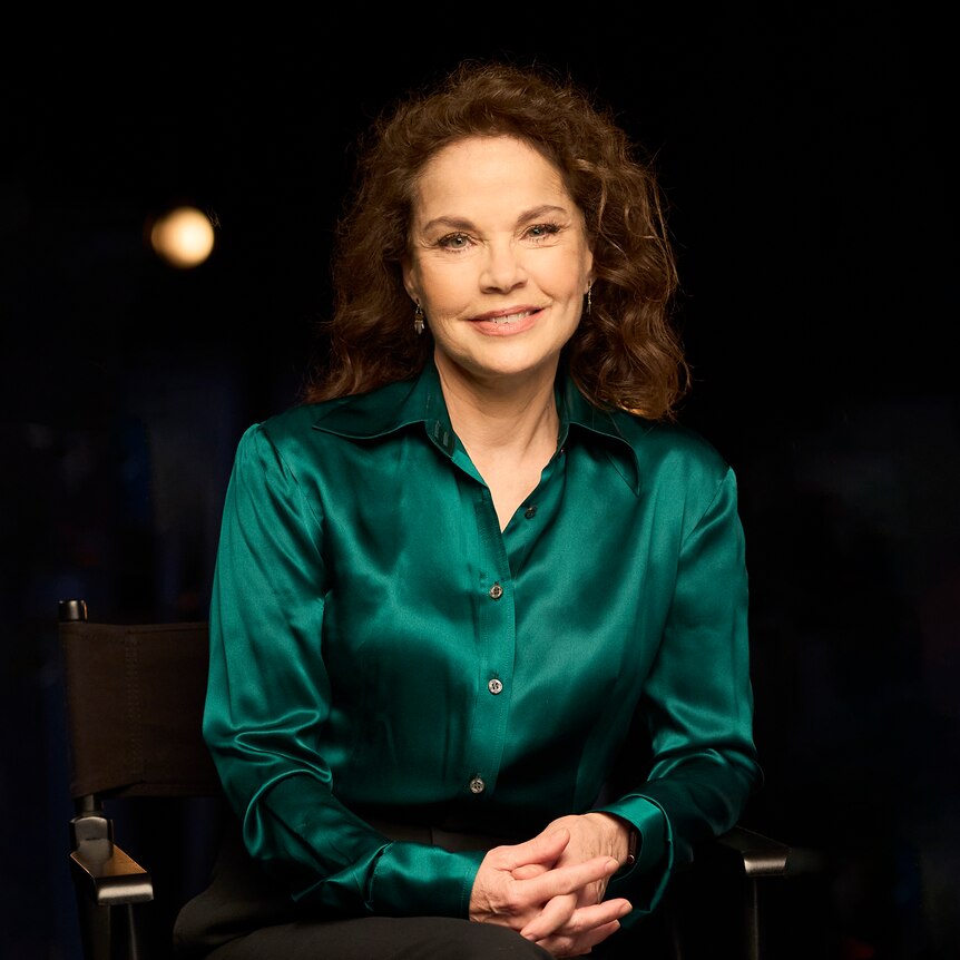 Actress Sigrid Thornton sits in a directors chair in a low-lit studio smiling.